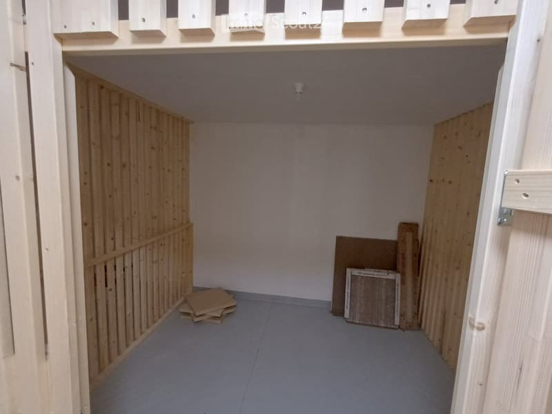 Empty room with wooden paneling on one wall, grey carpeted floor, and scattered wooden planks and boards.