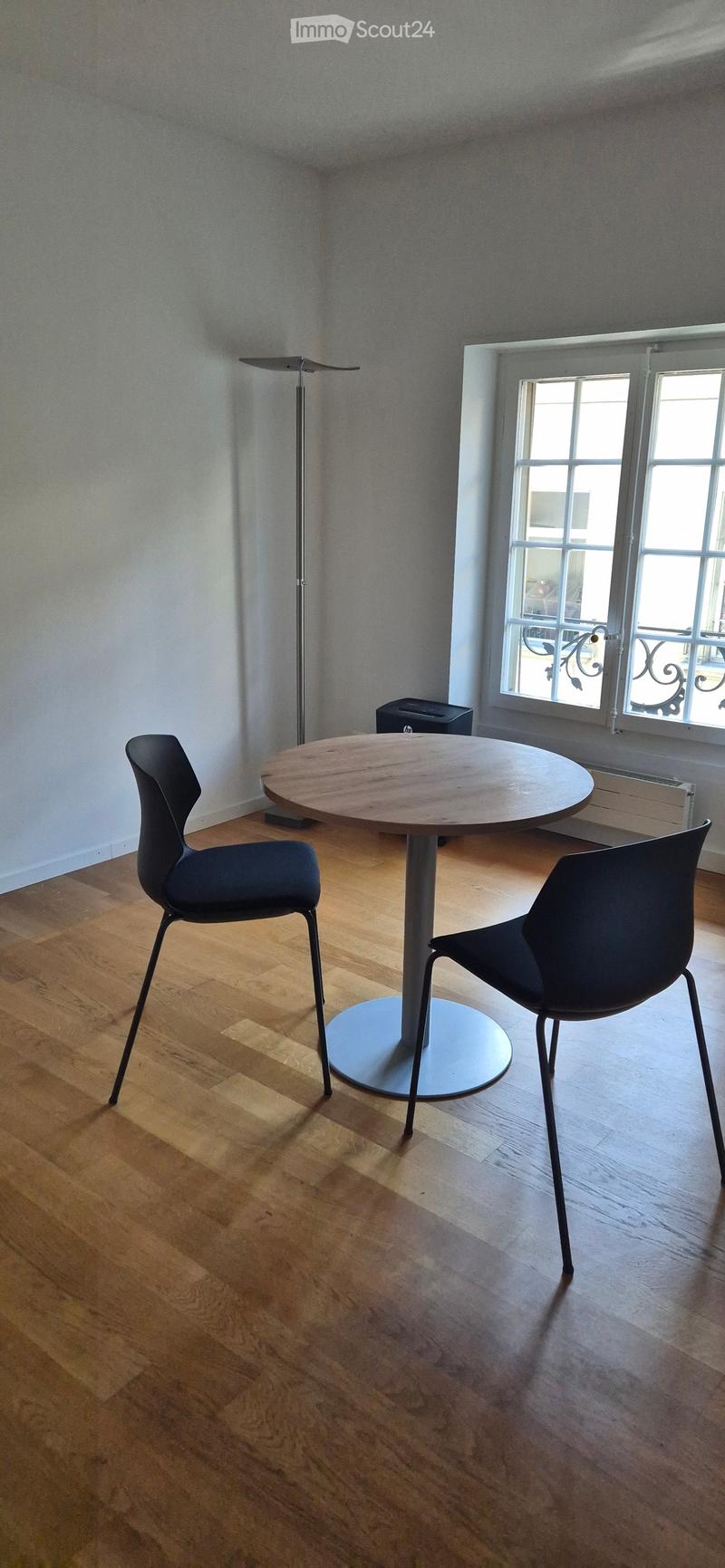 A small, modern room with wooden flooring, a round wooden table, two black chairs, and a floor lamp near a window with a view of a balcony.