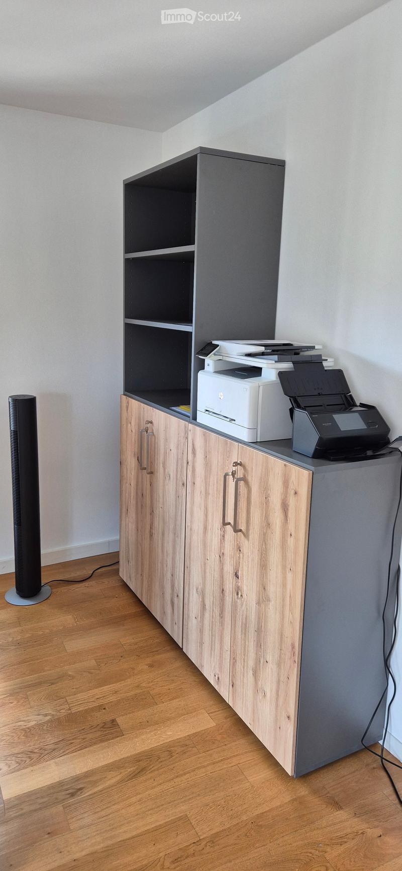 A small, modern office or workspace with a wooden cabinet, printer, and a standing fan. The room has light wooden flooring and white walls.