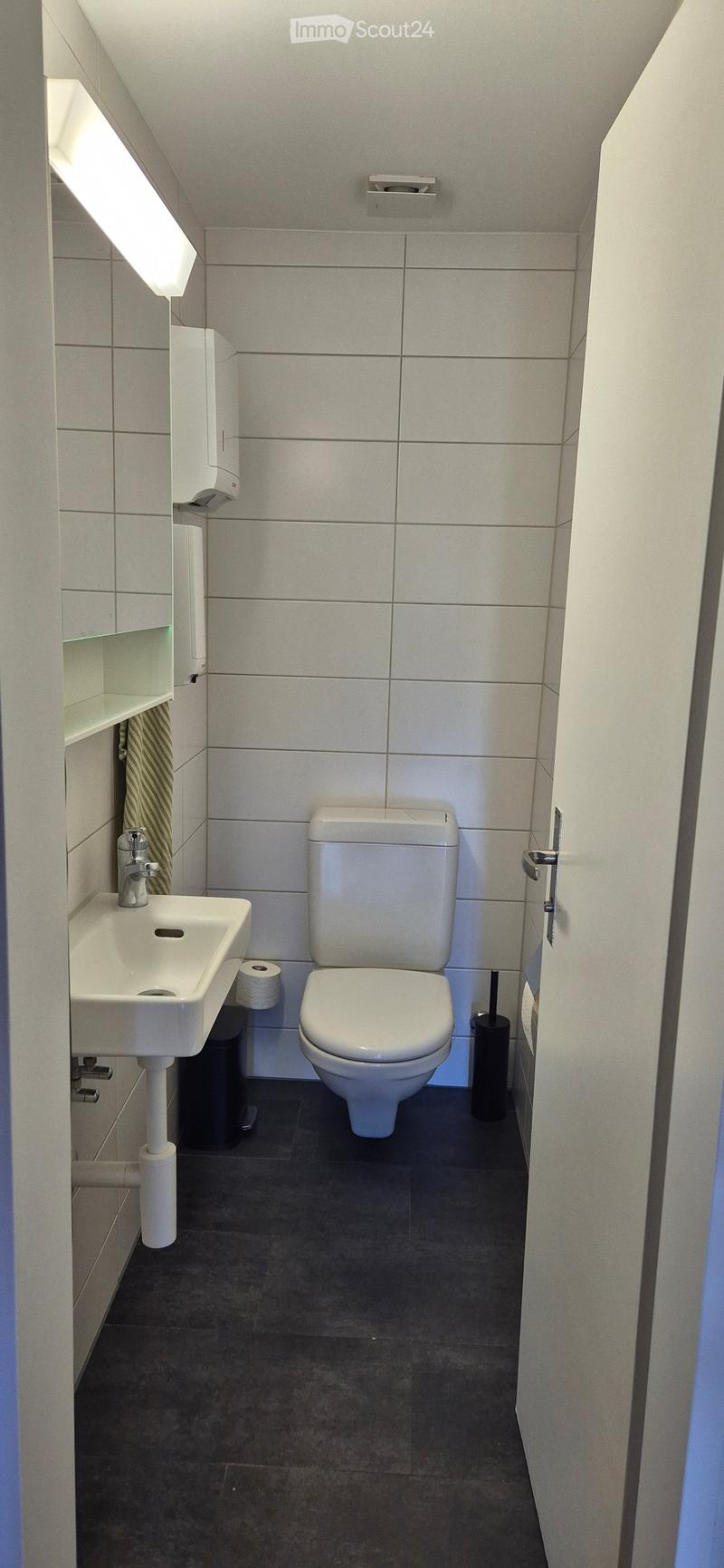 Modern bathroom with white tiles, a toilet, a sink with a chrome faucet, a hand dryer, and a mirror. The floor is dark grey.
