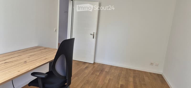 A small, unfurnished room with a wooden desk and a black office chair, white walls, and a closed white door.