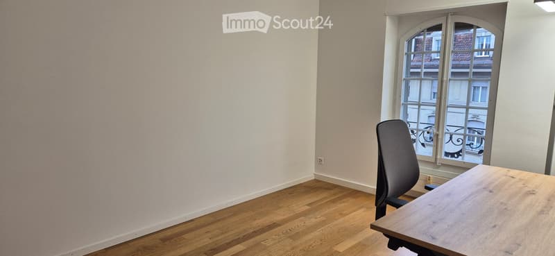 Empty room with wooden flooring, a single chair, and a table near a large window with a view of a building.