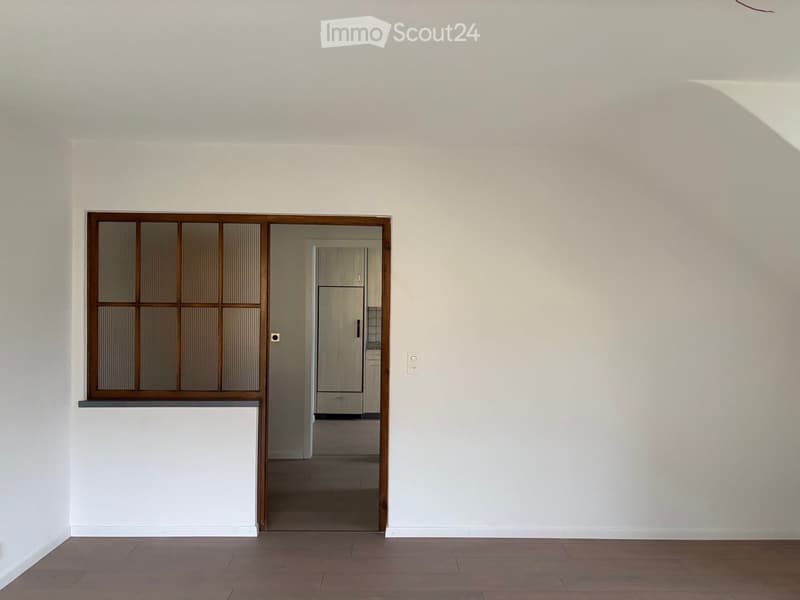 Empty room with light-colored walls, wooden flooring, and a doorway leading to another room with visible cabinets.