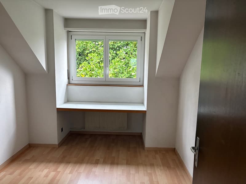 Empty room with wooden flooring, white walls, a window with a view of greenery, and a built-in bench beneath the window.