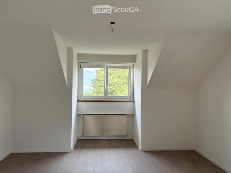 Empty room with wooden flooring, white walls, a window with a view of greenery, and a radiator below the window.