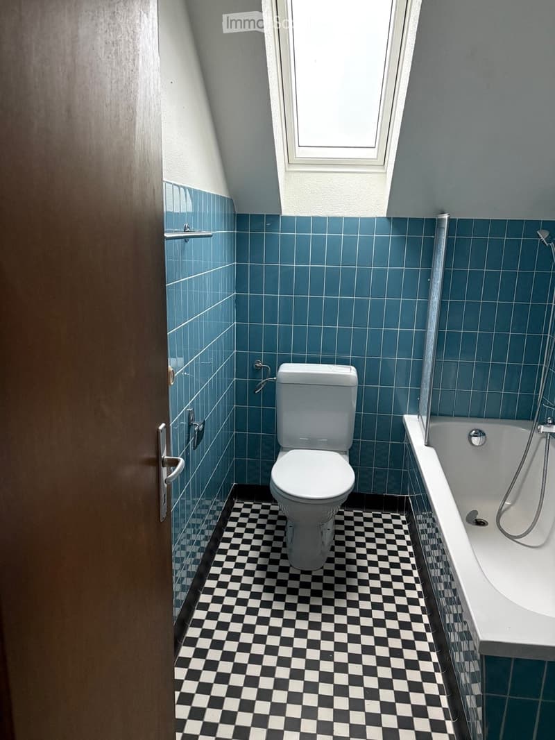 Bathroom with blue tiled walls, black and white checkered floor, toilet, bathtub, and a window above the bathtub.