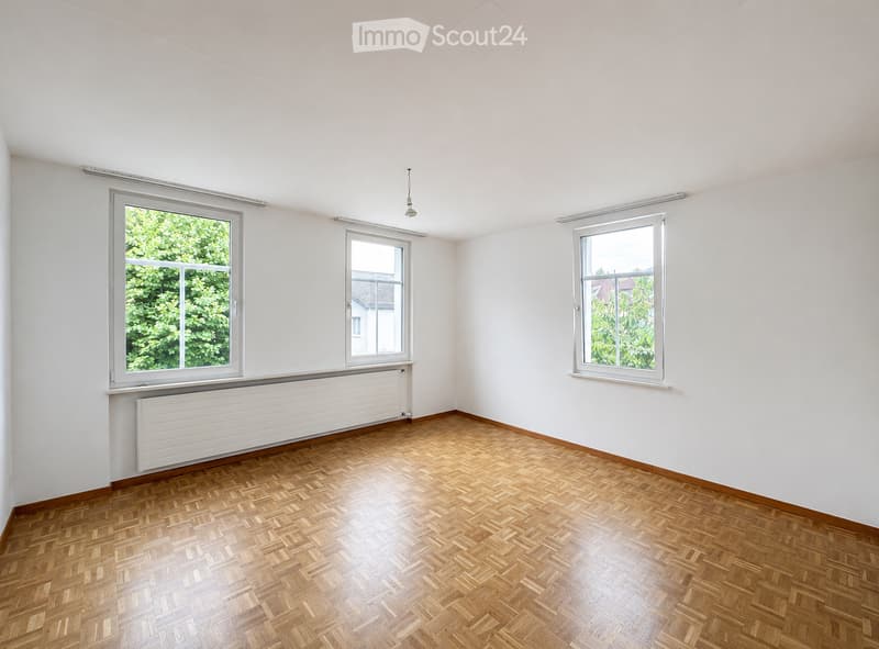 Empty room with white walls, wooden herringbone flooring, three windows, radiator, and a ceiling light fixture.