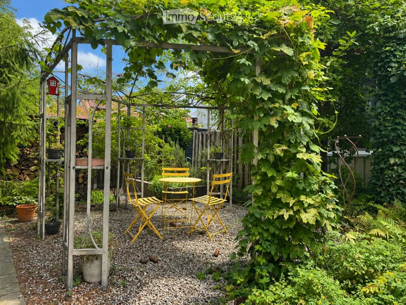 A lush garden with a pergola covered in vines, a small seating area with a yellow table and chairs, and various potted plants.
