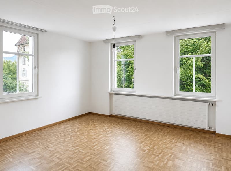 Empty room with white walls, wooden herringbone flooring, and three large windows offering views of greenery and a neighboring building. A radiator is visible beneath the center window, and a single light fixture hangs from the ceiling.