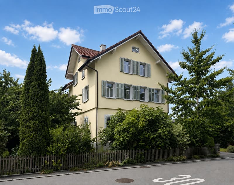 Two-story yellow house with white shutters and a red-tiled roof, surrounded by greenery and a wooden fence.