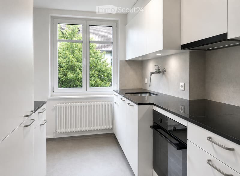 Modern kitchen with white cabinets, black countertops, stainless steel sink, built-in oven, and a window overlooking greenery.