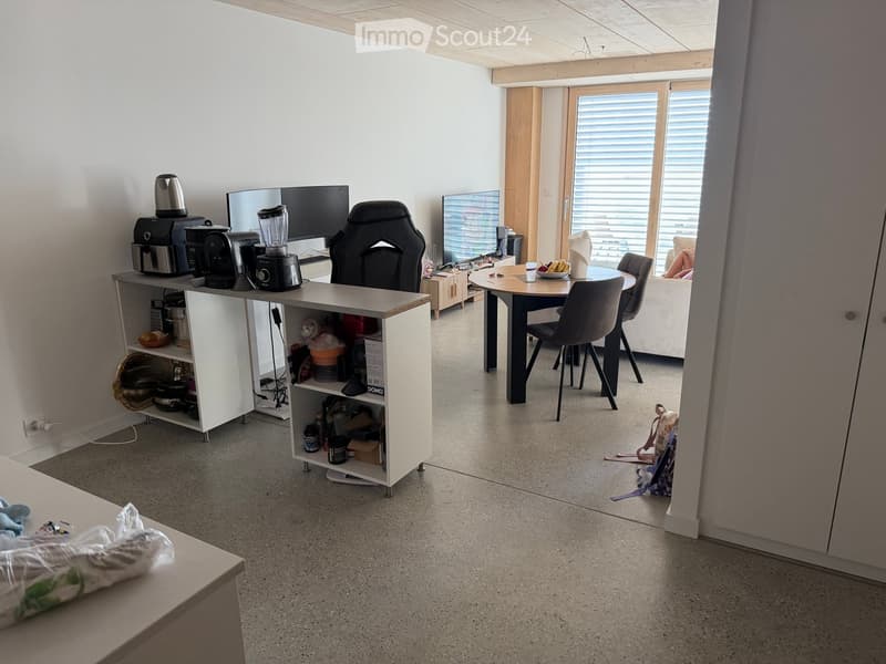 A modern, open-plan room with a kitchen area and a dining area. The kitchen features a coffee maker, blender, and a small cabinet. The dining area includes a round table with chairs and a screen. The room has large windows with blinds and a concrete floor