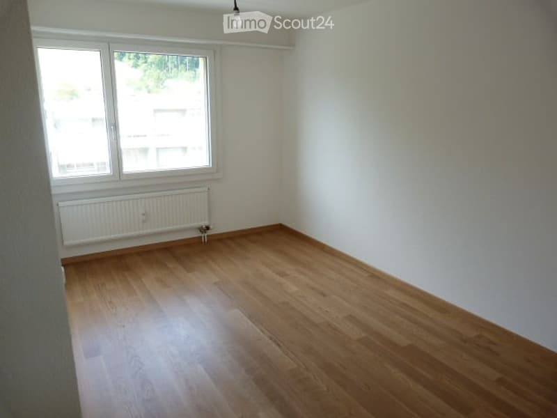 Empty room with wooden flooring, white walls, a window with white frame, and a radiator below the window.