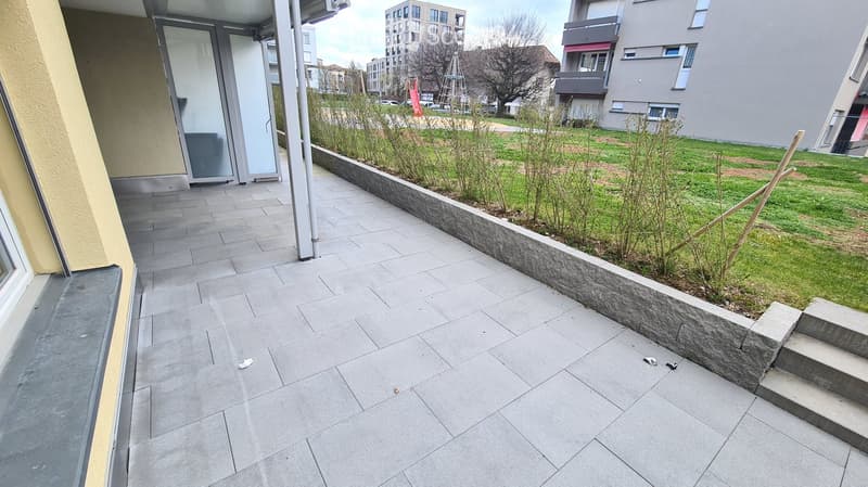 Balcony with grey tiled floor, low stone wall, small garden area with grass and plants, steps leading down, adjacent building with glass doors.