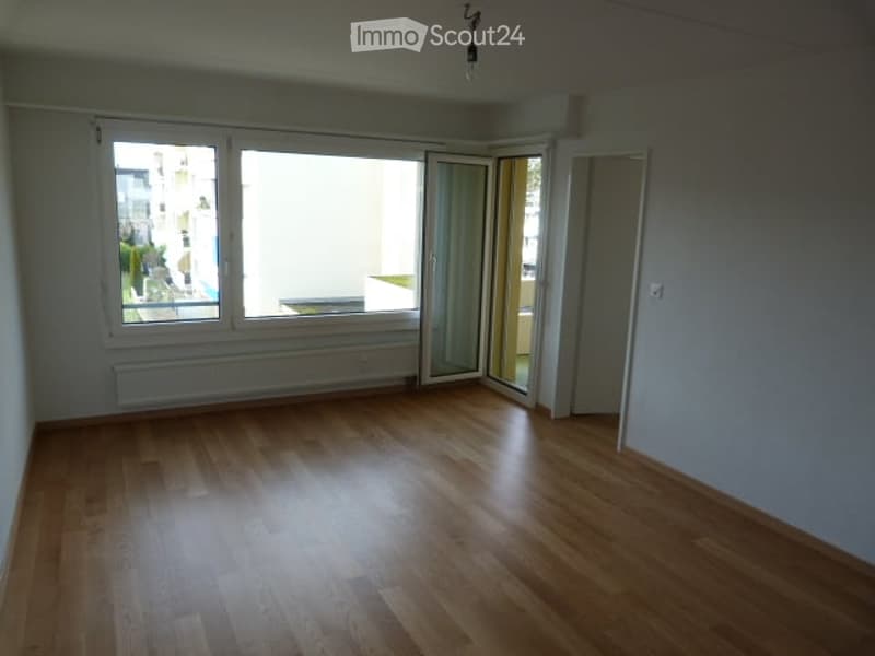 Empty room with wooden flooring, large windows, and a sliding glass door leading to a balcony.