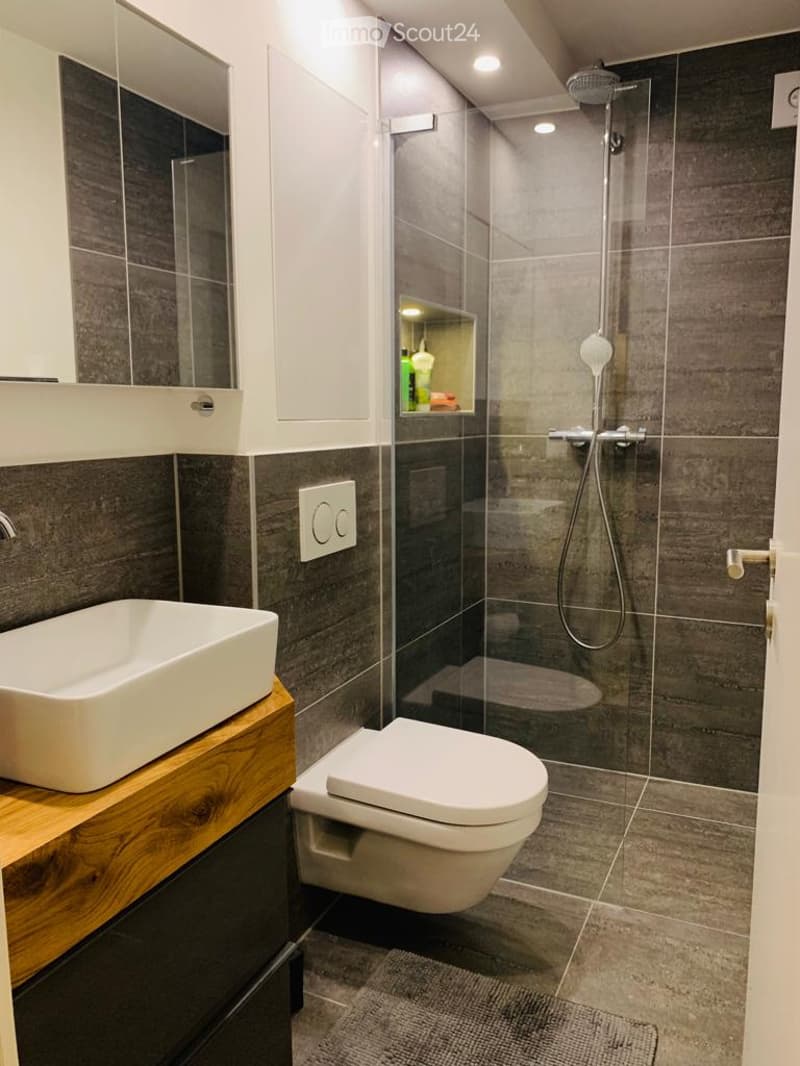 The image shows a modern bathroom with a glass-enclosed shower, a white toilet, and a wooden vanity with a vessel sink. The walls are tiled in a dark gray color, creating a sleek and contemporary look. The bathroom appears to be well-lit with recessed lig