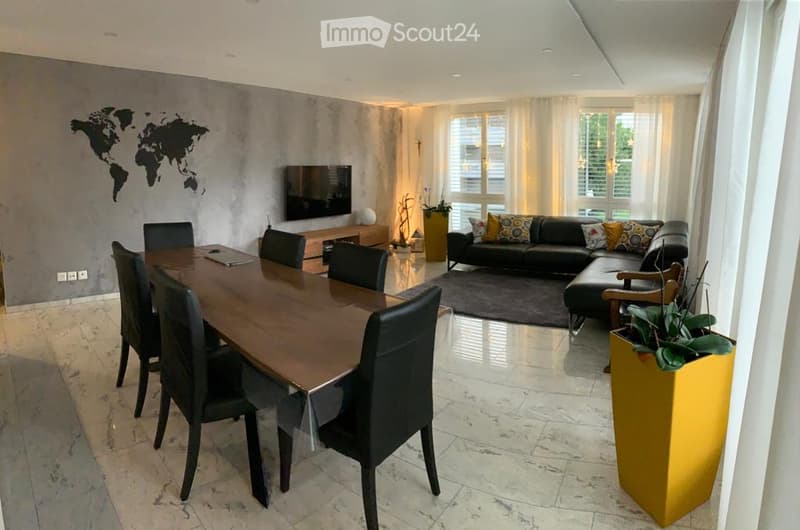 The image shows an open-plan living and dining area with a large dining table, black leather chairs, a sofa, and a TV mounted on the wall. The room has large windows that provide natural light and access to a balcony or terrace. The floor is covered in ma