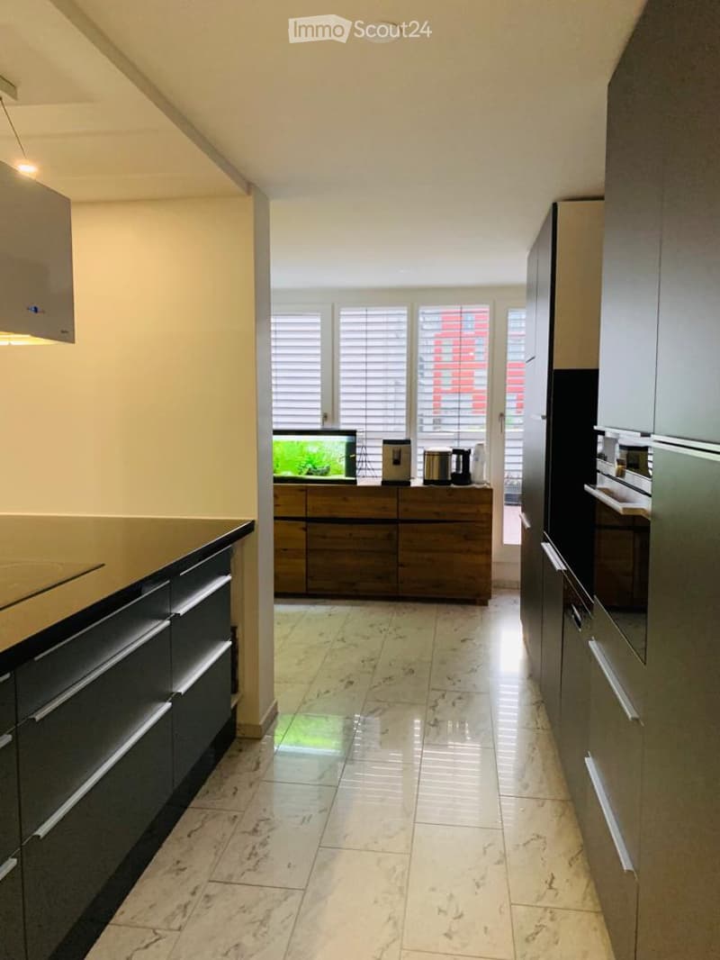 Spacious kitchen with marble tile flooring, black cabinets, wooden drawers, and a dishwasher visible on the counter. Large windows provide natural light and a view of the exterior.