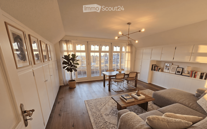 Modern living and dining area with a grey sectional sofa, coffee table with books, dining table with four chairs, large windows with sheer curtains, built-in white cabinets, and a potted plant.
