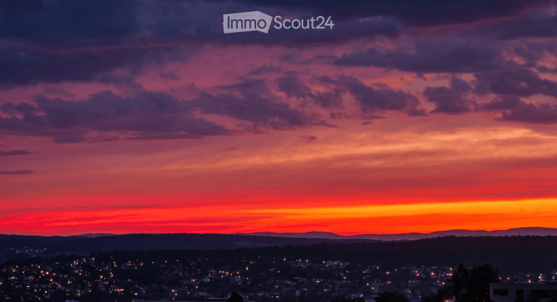Panoramic view of a cityscape at sunset, with vibrant red and orange hues in the sky and scattered lights from buildings below.