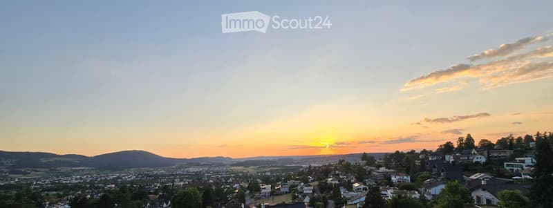 Panoramic view of a suburban neighborhood at sunset, with houses, trees, and distant hills.