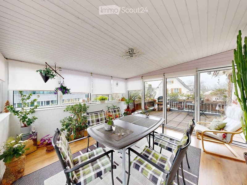 A covered balcony with a wooden floor, a dining table with four chairs, potted plants, and a view of the outdoor area.