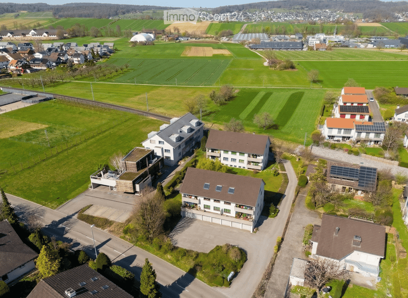 Aerial view of a residential area with several houses, green fields, and solar panels on the roofs. The houses have different architectural styles, some with balconies and terraces. The area is surrounded by trees and bushes, and there is a swimming pool