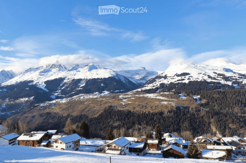 Group of chalets with snowy roofs nestled in a mountain valley surrounded by pine trees and snow-capped mountains.