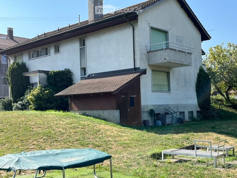 Two-story house with white exterior, multiple windows, and a balcony on the upper floor. A wooden garage is attached to the side, and the front yard includes a trampoline and outdoor furniture.