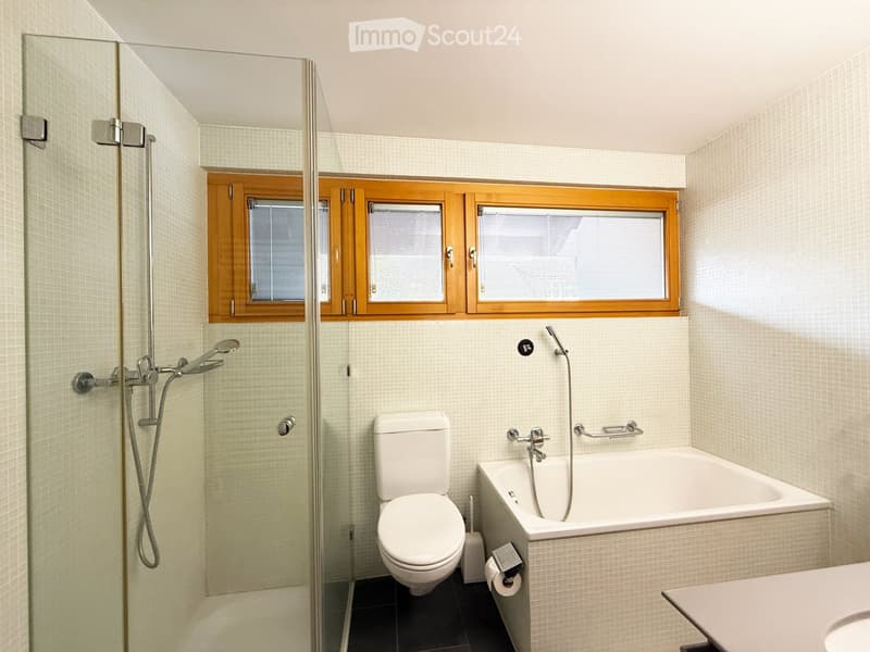 Modern bathroom with a bathtub, toilet, shower, and a window with wooden frame above the bathtub.