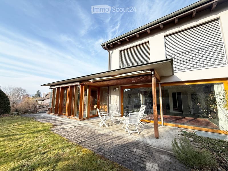 A modern house with a spacious outdoor patio area featuring white chairs and a table, surrounded by a well-maintained lawn and paved pathways.