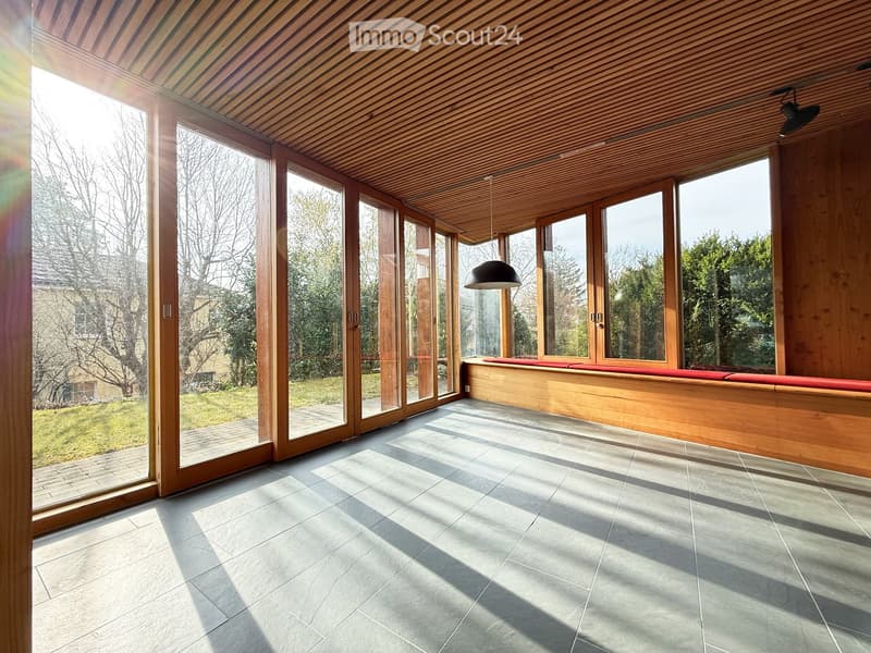 Bright, wooden-framed room with large glass doors leading to a garden, featuring a built-in wooden bench with red cushions and a hanging pendant light.