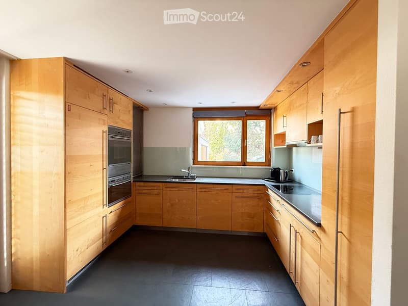 Modern kitchen with light wooden cabinets, stainless steel oven, sink, and countertop, featuring a large window with a view of greenery.