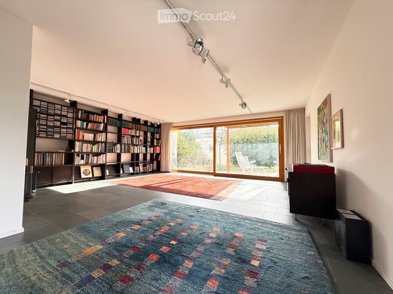 Bright living room with large windows, bookshelves, colorful rugs, and modern lighting fixtures.