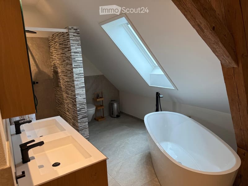 Modern bathroom with a large bathtub, double sink, sleek faucets, and a skylight window. Features a wooden shelf and a small trash can.