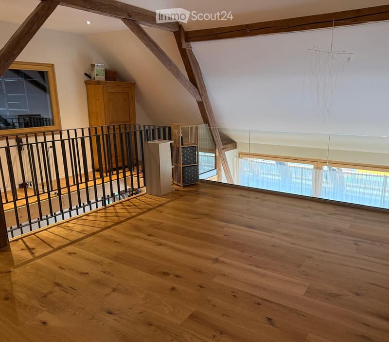 Bright, open attic space with wooden floors, exposed wooden beams, and a modern glass railing overlooking a lower level. The room is empty, with no furniture or furnishings visible.