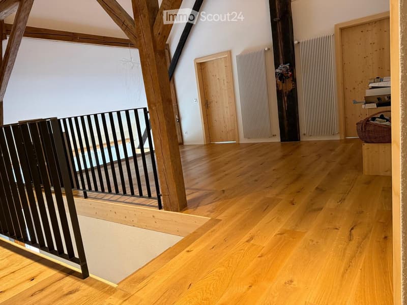 Spacious hallway with wooden flooring, exposed wooden beams, and a black metal railing leading to a lower level. Features include white walls, wooden doors, and radiators.
