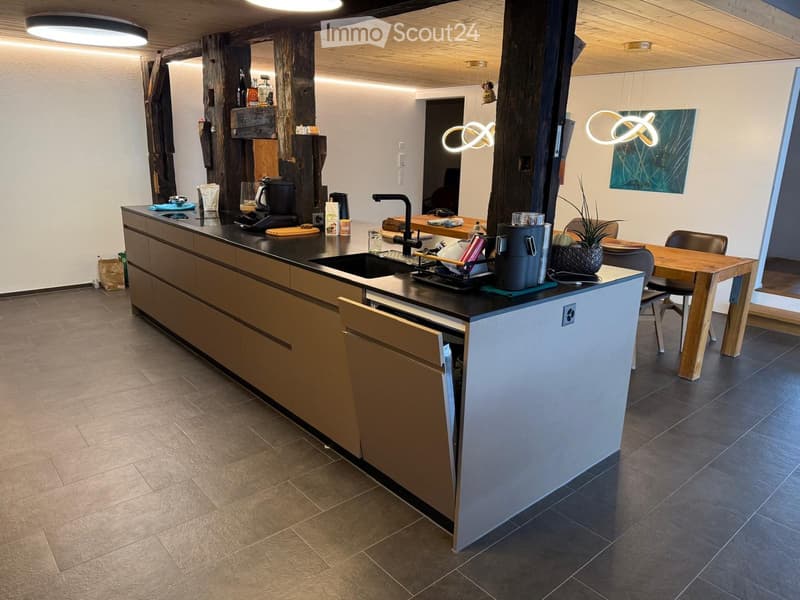Modern kitchen with a central island, dark countertops, and wooden cabinets. Includes a sink, coffee maker, dish rack, blender, and a small dining area with a table and chairs.