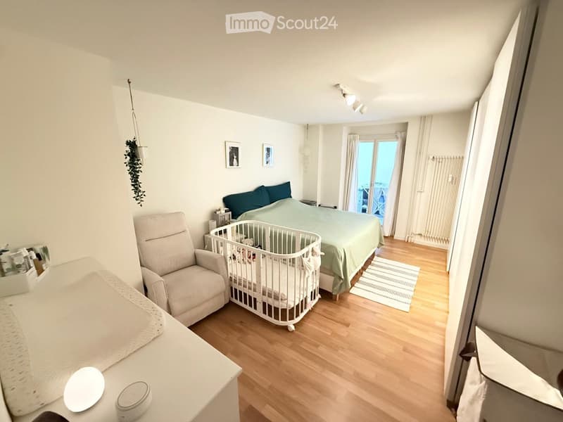 Bright bedroom with wooden flooring, a large bed with green bedding, a white crib, a beige armchair, a small table with decorative items, and a view through a large window with curtains.