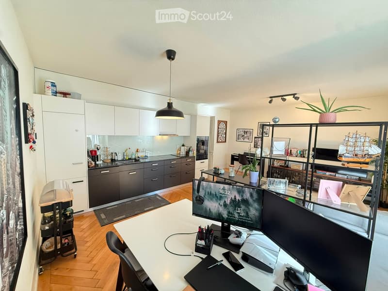 A modern, open-plan living and working space with a kitchen area, a desk setup, and a shelf with decorative items. The room features light wood flooring, white cabinets, and a sleek black desk with dual monitors.