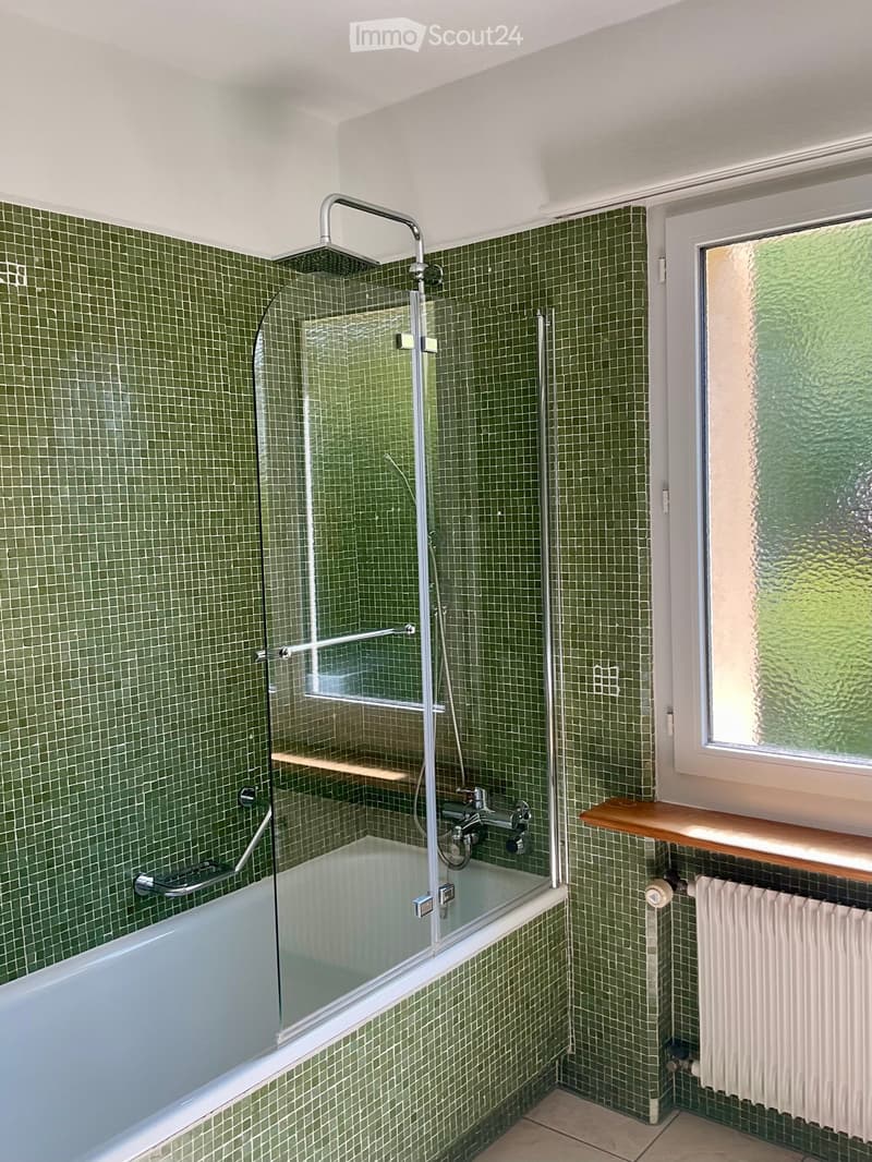 Bathroom with green tiled walls, a bathtub with a glass shower enclosure, a wooden shelf, and a radiator.