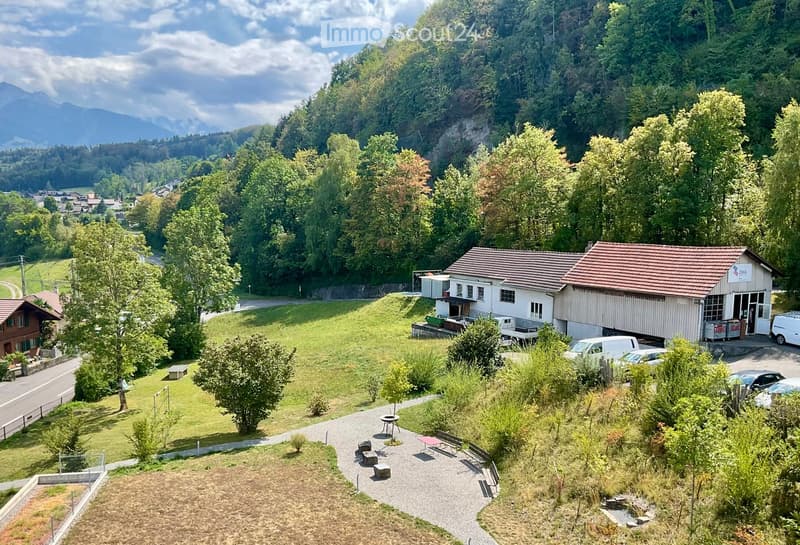 Aerial view of a rural building complex with a red-tiled roof, surrounded by lush greenery and trees, with a parking area and outdoor seating area.