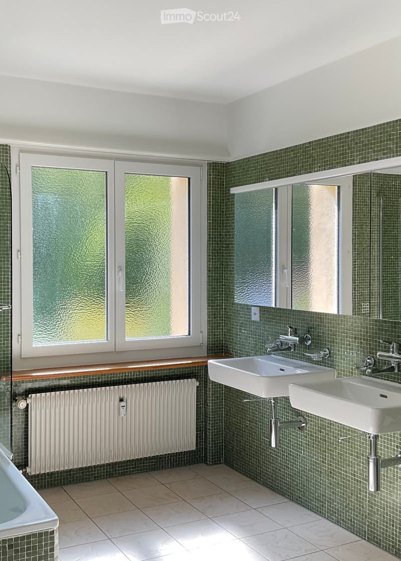 Bathroom with green tiled walls, two sinks, a bathtub, and a large mirror above the sinks.