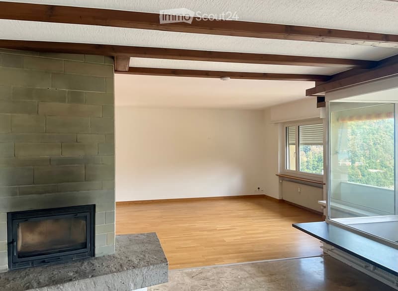 Empty room with wooden flooring, exposed wooden beams, fireplace, large windows, and a view of greenery.