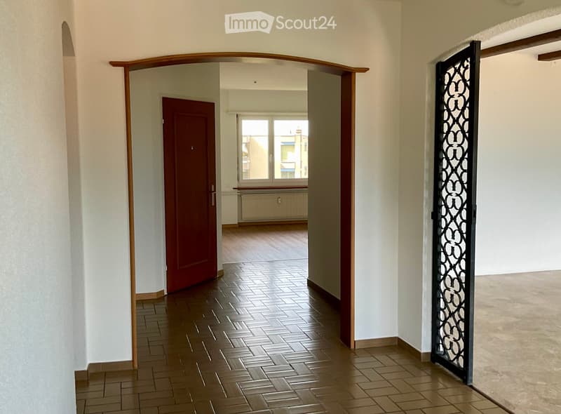 Empty hallway with tiled floor, white walls, wooden archway, red door, and black decorative gate leading to another room with wooden flooring and a window.