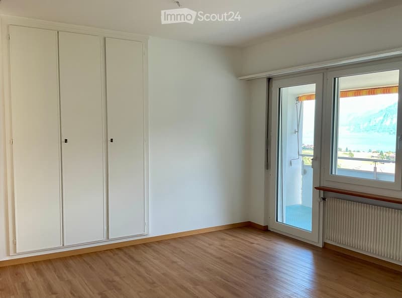 Empty room with wooden flooring, white walls, large sliding glass door leading to a balcony, and a window with a scenic view.