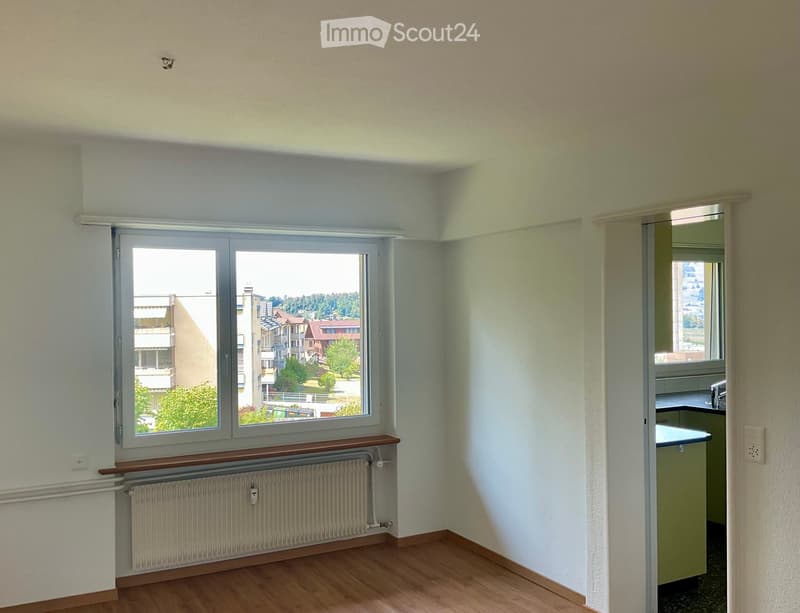 Empty room with wooden flooring, large window with view of buildings and greenery, radiator, and a small kitchen area visible through an open doorway.