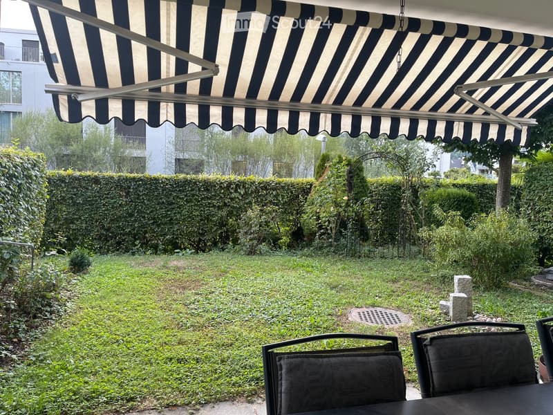 Garden with a covered patio area, black chairs and table, surrounded by greenery and a hedge.