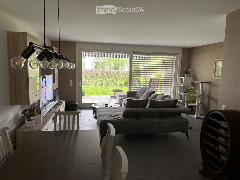 Modern living room with a grey sectional sofa, coffee table, dining table, chairs, cat tree, potted plants, and a wine rack. Large windows and sliding doors lead to a patio area.