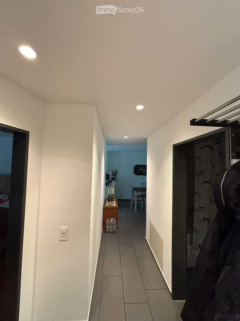A narrow hallway with white walls, tiled floor, and recessed ceiling lights. Doors lead to adjacent rooms, including a visible dining area with a table and chairs.
